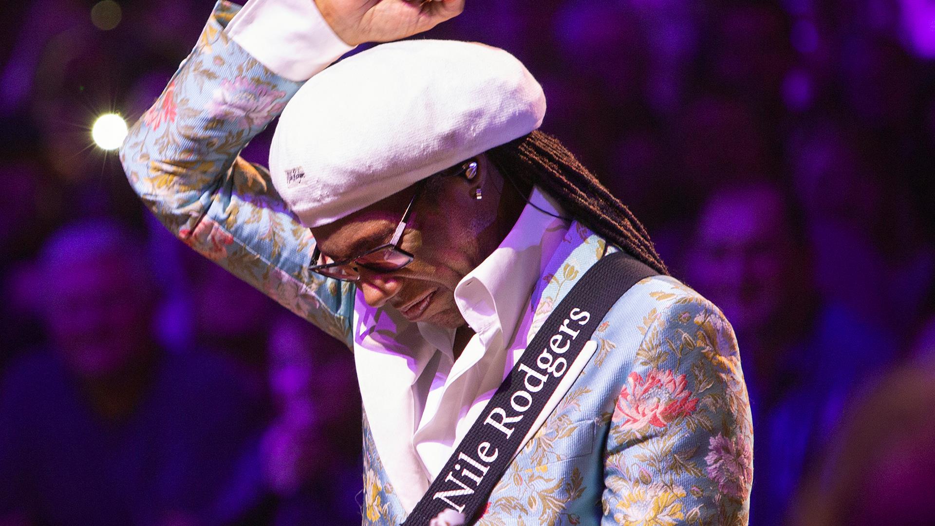Nile Rodgers in floral blue blazer and white beret, lifts arm above head when performing for crowd.